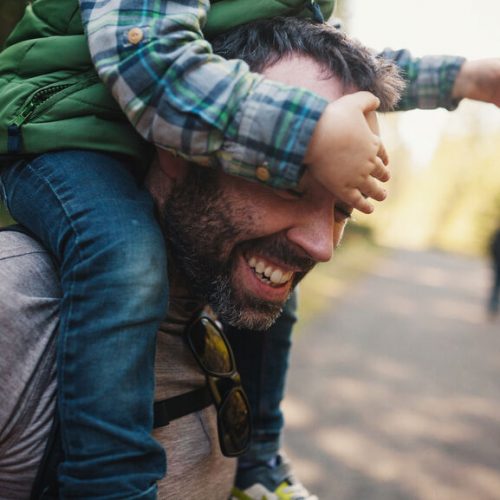 Young Dad Laughing Outside With Little Boy On His Shoulders Blindfolding Him